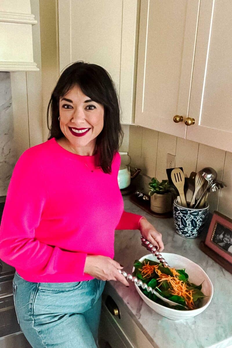 Person in a bright pink sweater holds salad tongs over a bowl of greens and shredded carrots in a kitchen. Utensils and a framed photo are on the counter.