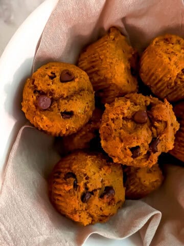 Eight pumpkin muffins in a white bowl with a tan napkin on a marble counter. Chocolate chips are sprinkled around and there is a small bowl of salt next to the bowl of muffins.