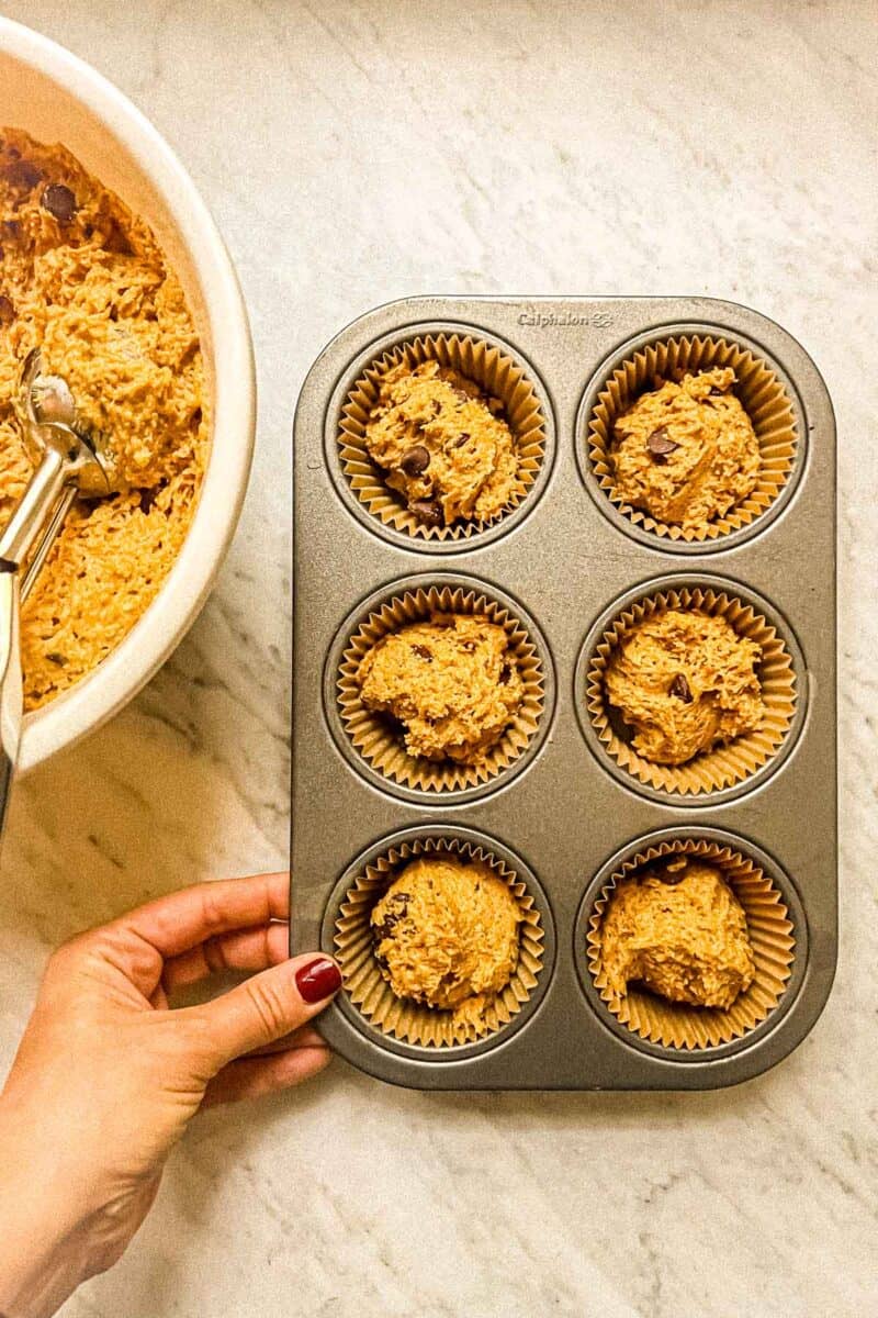 A muffin baking pan filled with batter sitting on a counter. The bowl of batter is sitting to the left of the muffin pan.