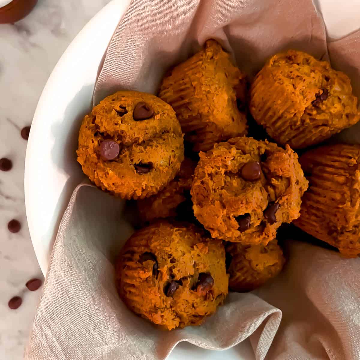 Eight Kodiak pumpkin muffins in a white bowl with a tan napkin on a marble counter. Chocolate chips are sprinkled around and there is a small bowl of salt next to the bowl of muffins.