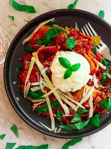 Saturday Night Tofu Bolognese in a bowl on a marble counter. Surrounded by basil leaves and a glass of water.