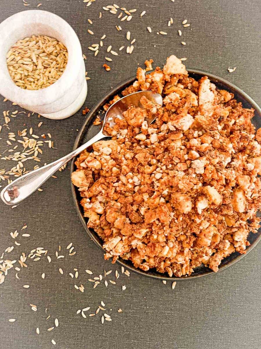 Prepared Italian Sausage Tofu Crumbles in a bowl with a spoon on the countertop. Bowl is surrounded by fennel seeds on the counter and a bowl of additional seeds.