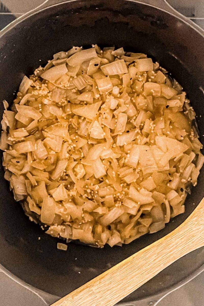 Onions and garlic sauteing in a cast iron Dutch oven on a stovetop. A wooden spoon is resting on the side of the Dutch oven.