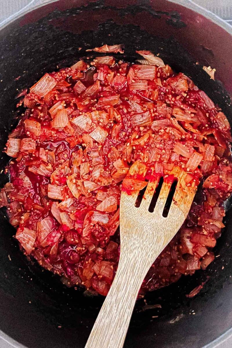 Onion, garlic, tomato paste, oregano, nutmeg, salt, and pepper are in a cast iron Dutch oven. A wooden spoon is resting on the pot.