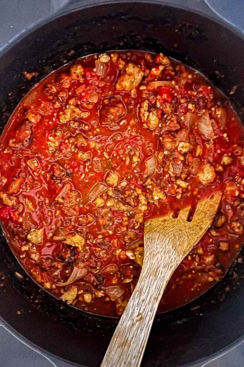 Onion, garlic, tomato paste, oregano, nutmeg, salt, pepper marinara, Extra Virgin Olive Oil, chicken broth and tofu crumbles are in a cast iron Dutch oven. A wooden spoon is resting on the pot.