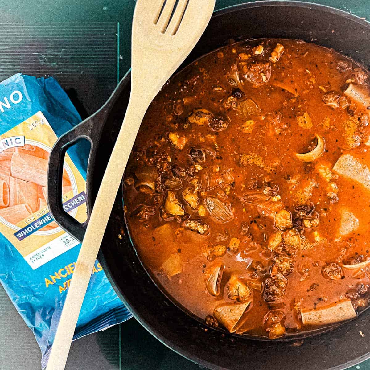 Onion, garlic, tomato paste, oregano, nutmeg, salt, pepper marinara, Extra Virgin Olive Oil, chicken broth and tofu crumbles, and whole wheat noodles are in a cast iron Dutch oven. A wooden spoon is resting on the pot and a bag of whole wheat pasta is next to it.