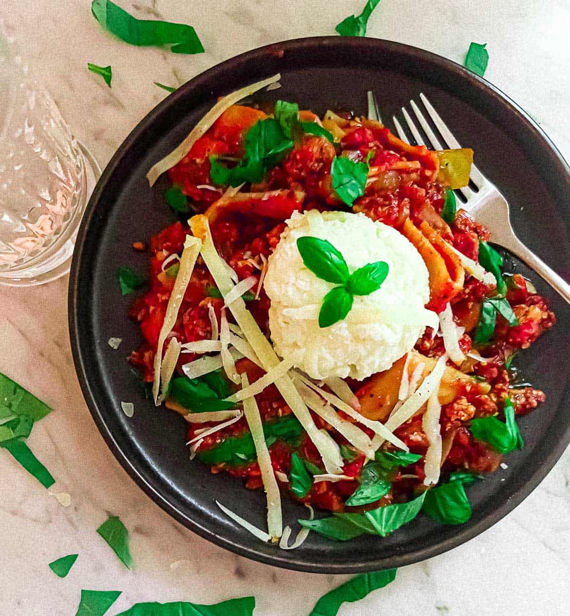 Saturday Night Tofu Bolognese in a bowl on a marble counter. Surrounded by basil leaves and a glass of water.