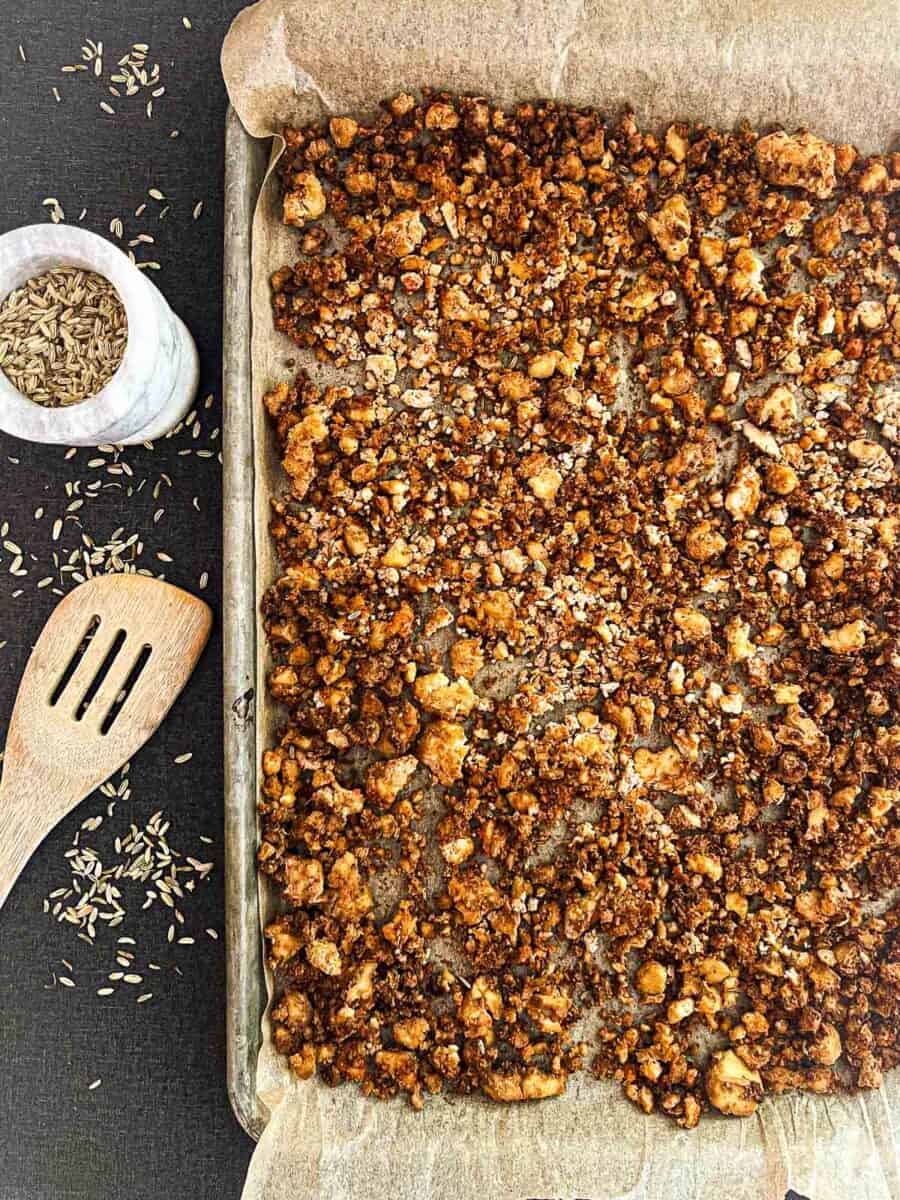 Baked sausage crumbles on a cookie sheet lined with parchment paper. Fennel seeds and wooden spoon are on the left of the baking sheet.