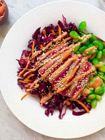 Seared ahi tuna salad in a bowl on countertop. Sesame seeds, carrot shreds, and a glass of water surround it.