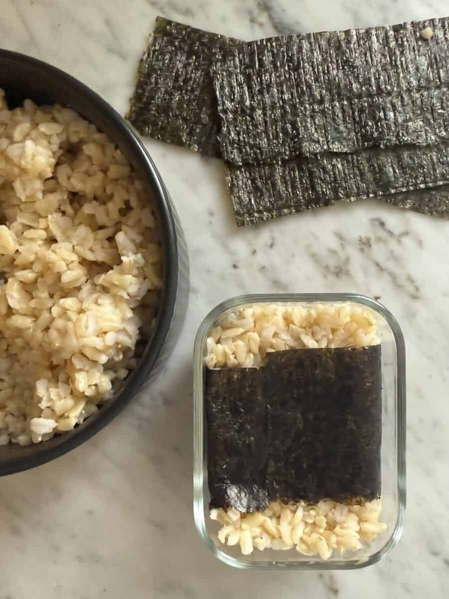A salmon musubi in the mold with a bowl of rice and nori strips beside it.