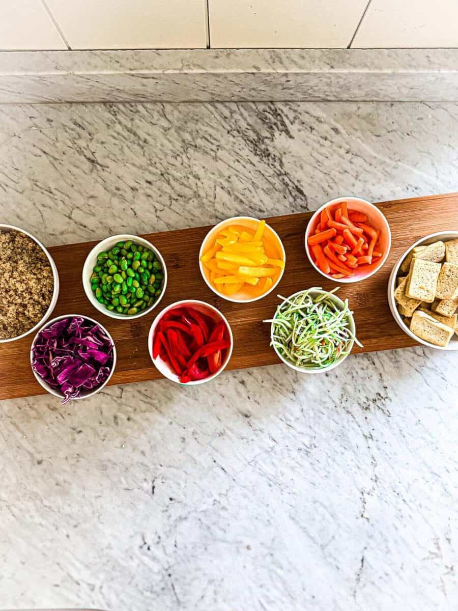 Bowls of prepared veggies, quinoa, and tofu on a board