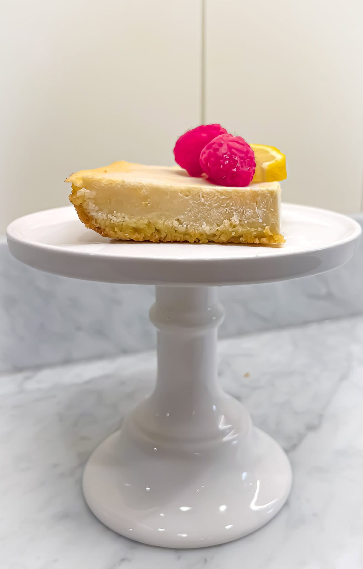 A Protein Lemon Bar garnished with raspberries and a lemon wedge on a white pedestal serving dish.