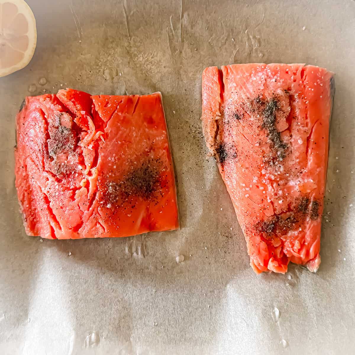 Two salmon filets prepared with salt and pepper on a parchment covered baking sheet with a lemon slice on the side.