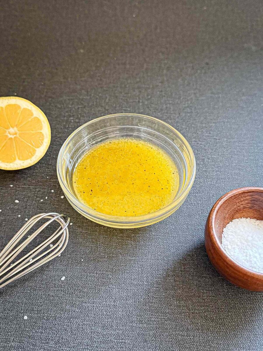 Lemon vinaigrette in a small bowl with whisk, salt and lemon to the side.