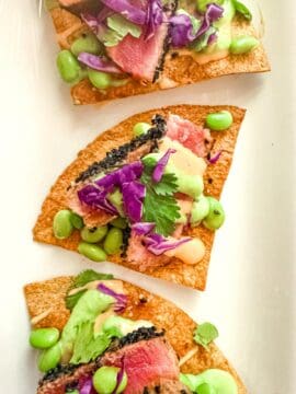 Ahi tuna nachos topped with seared tuna, purple cabbage, edamame, cilantro, horseradish sauce, and spicy mayo arranged on a white plate. Fresh and vibrant.