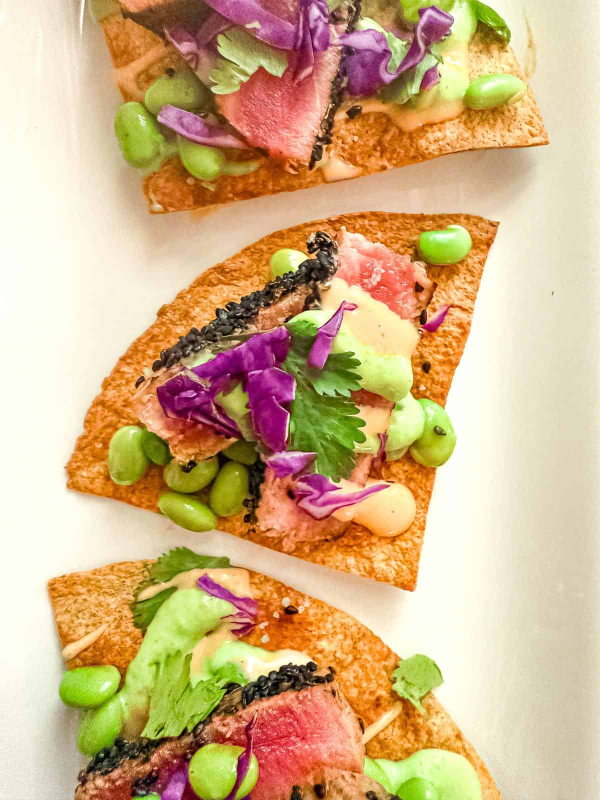 Ahi tuna nachos topped with seared tuna, purple cabbage, edamame, cilantro, horseradish sauce, and spicy mayo arranged on a white plate. Fresh and vibrant.