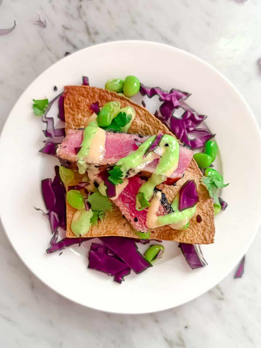 An ahi tuna nacho topped with seared tuna, purple cabbage, edamame, cilantro, horseradish sauce, and spicy mayo arranged on a white plate. Fresh and vibrant.