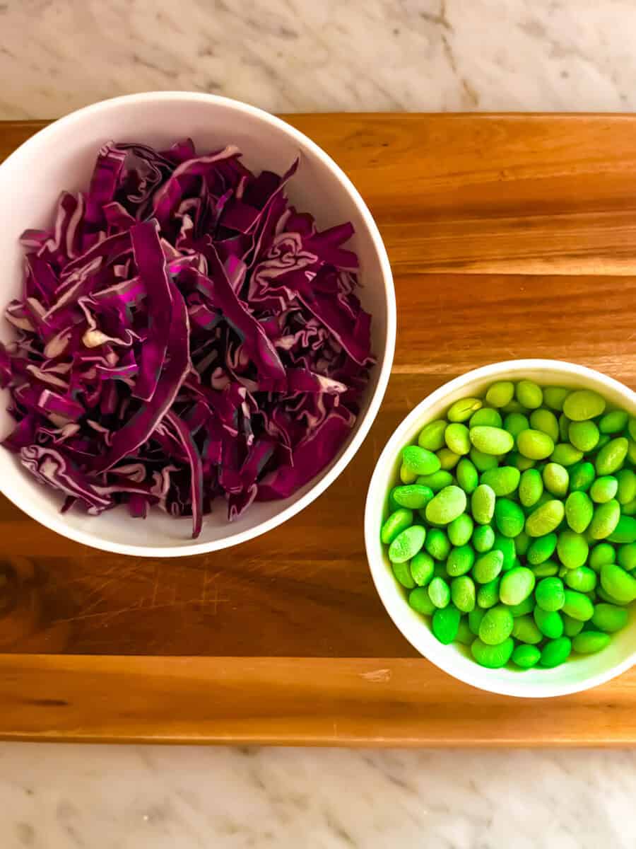 Two bowls of garnishes for ahi tuna nachos resting on a cutting board atop a marble counter. One bowl contains shredded red cabbage and the other contains prepared edamame.