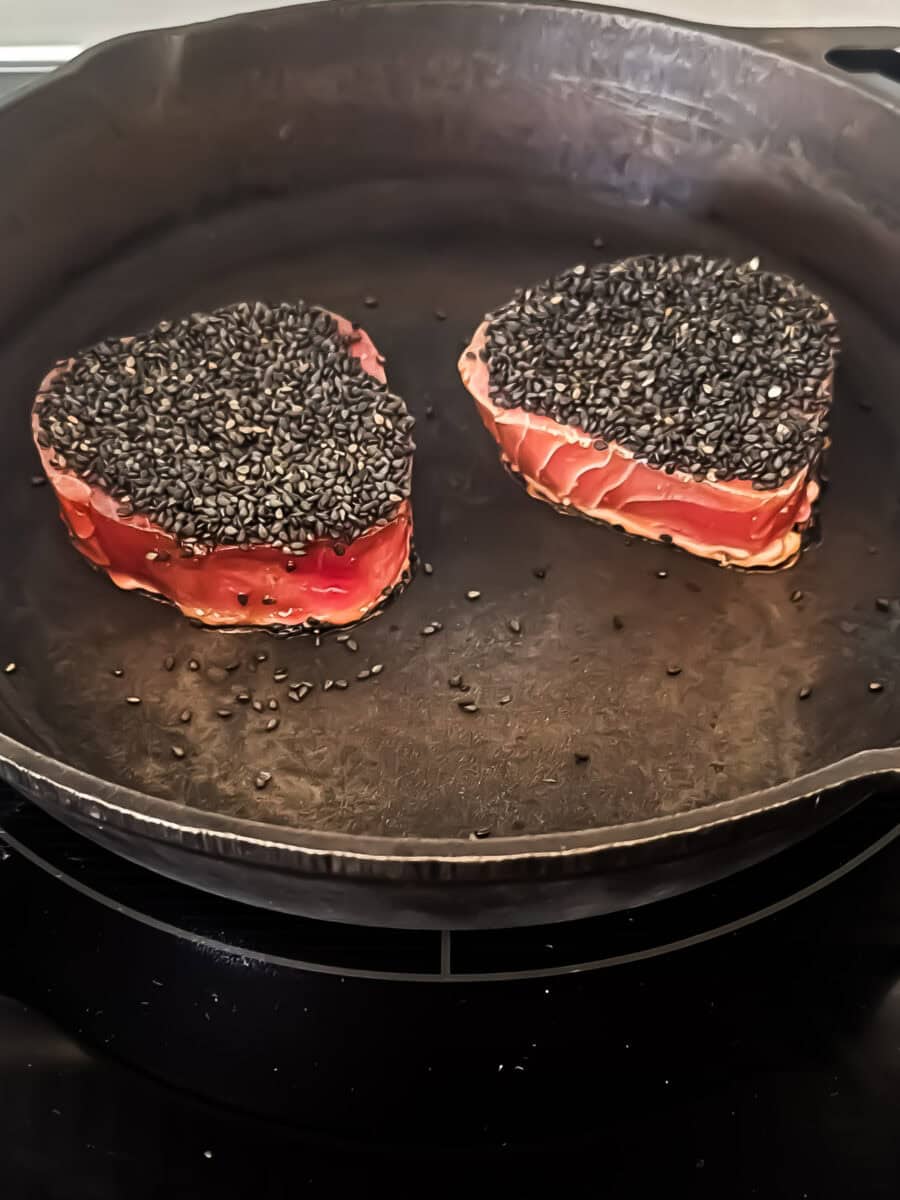 Two ahi tuna filets encrusted in black sesame seeds are cooking in a cast iron pan on a stove.