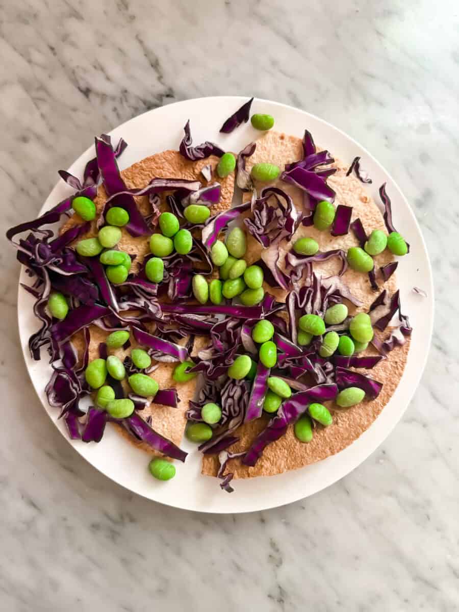 Whole wheat tortillas topped with shredded red cabbage and prepared edamame on a white plate.