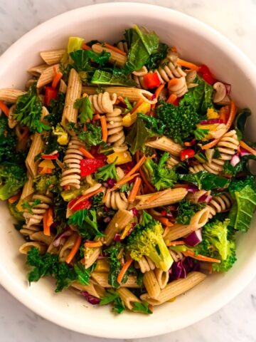 A vibrant kale pasta salad in a white bowl, featuring whole wheat rotini, penne, kale, broccoli, carrots, red peppers, yellow peppers, corn, purple cabbage and parsley on a marble background.