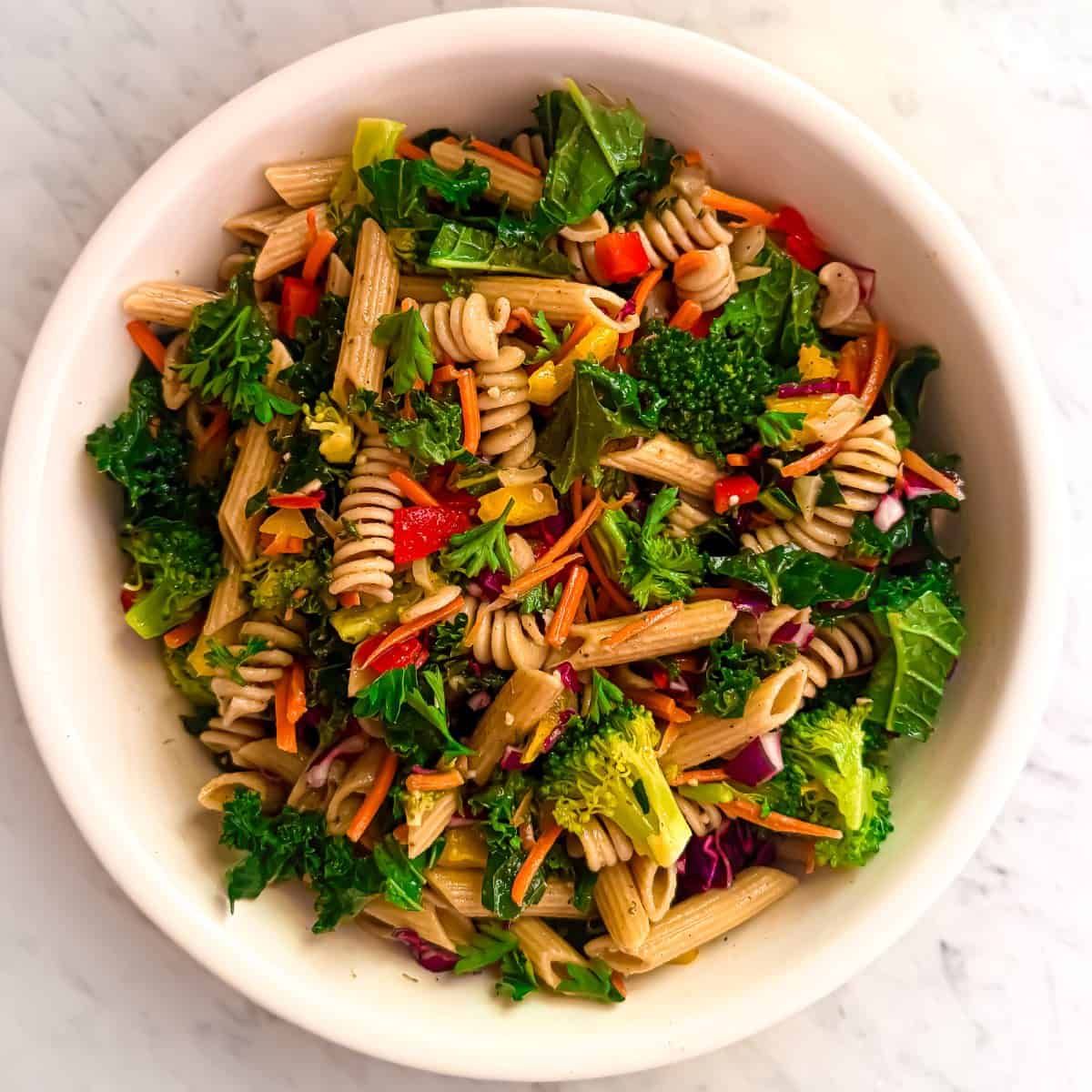 A vibrant kale pasta salad in a white bowl, featuring whole wheat rotini, penne, kale, broccoli, carrots, red peppers, yellow peppers, corn, purple cabbage and parsley on a marble background.