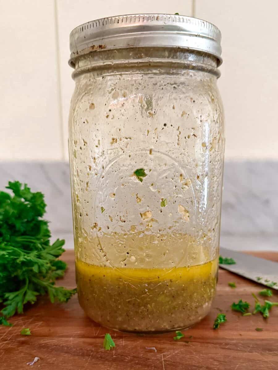 A quart Mason jar on a counter with parsley and a knife. The Mason jar contains a dressing with EVOO, parsley, and garlic visible.