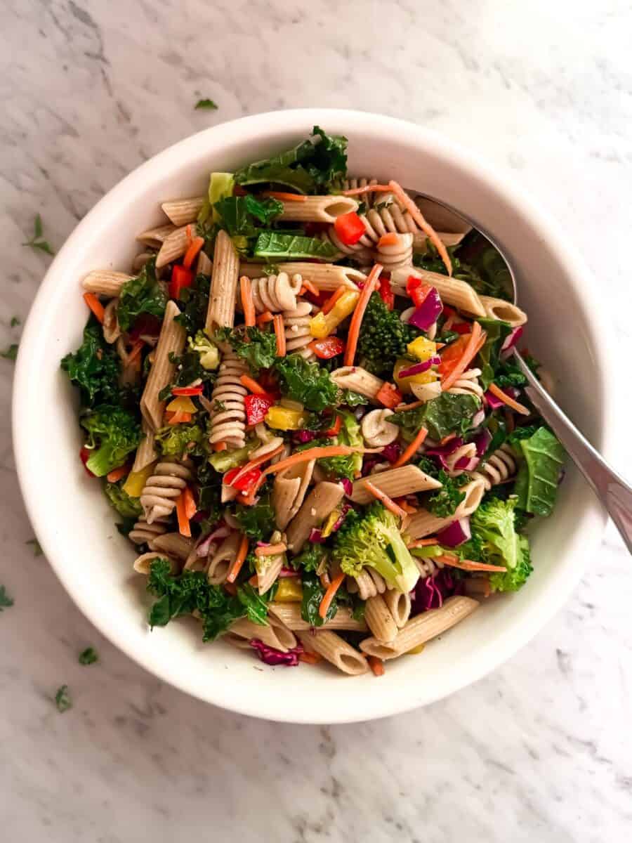 A vibrant pasta salad in a white bowl with a serving spoon, featuring whole wheat rotini, penne, kale, broccoli, carrots, red peppers, yellow peppers, corn, purple cabbage and parsley on a marble background.