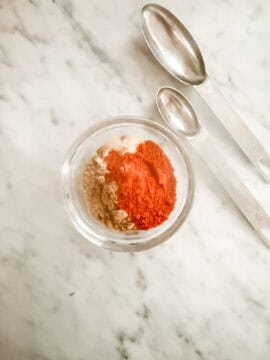 Ingredients for low sodium fajita seasoning in a class bowl flanked by two measuring spoons all on a marble countertop.