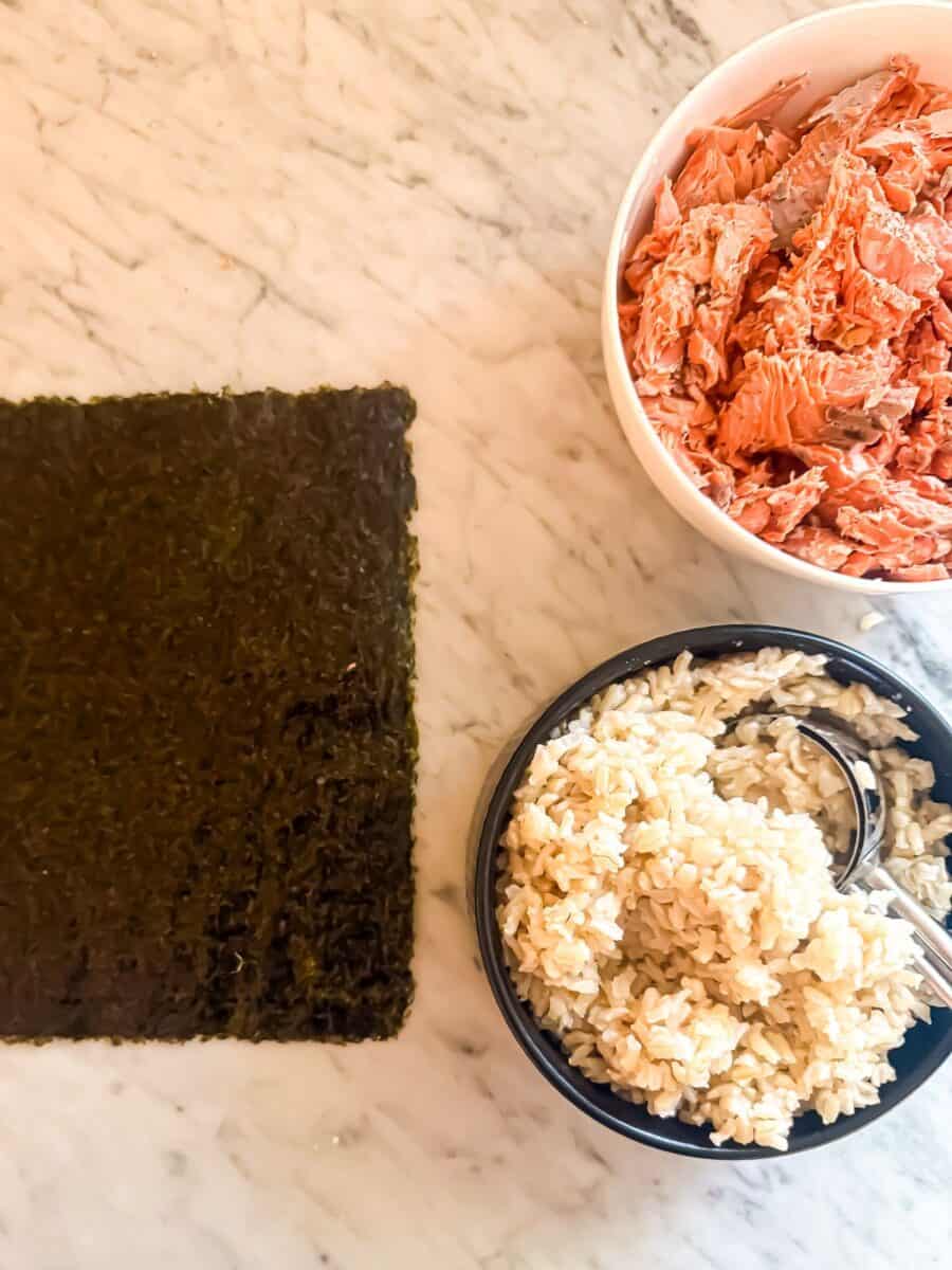 Shredded salmon and brown rice in separate bowls with a sheet of nori beside them