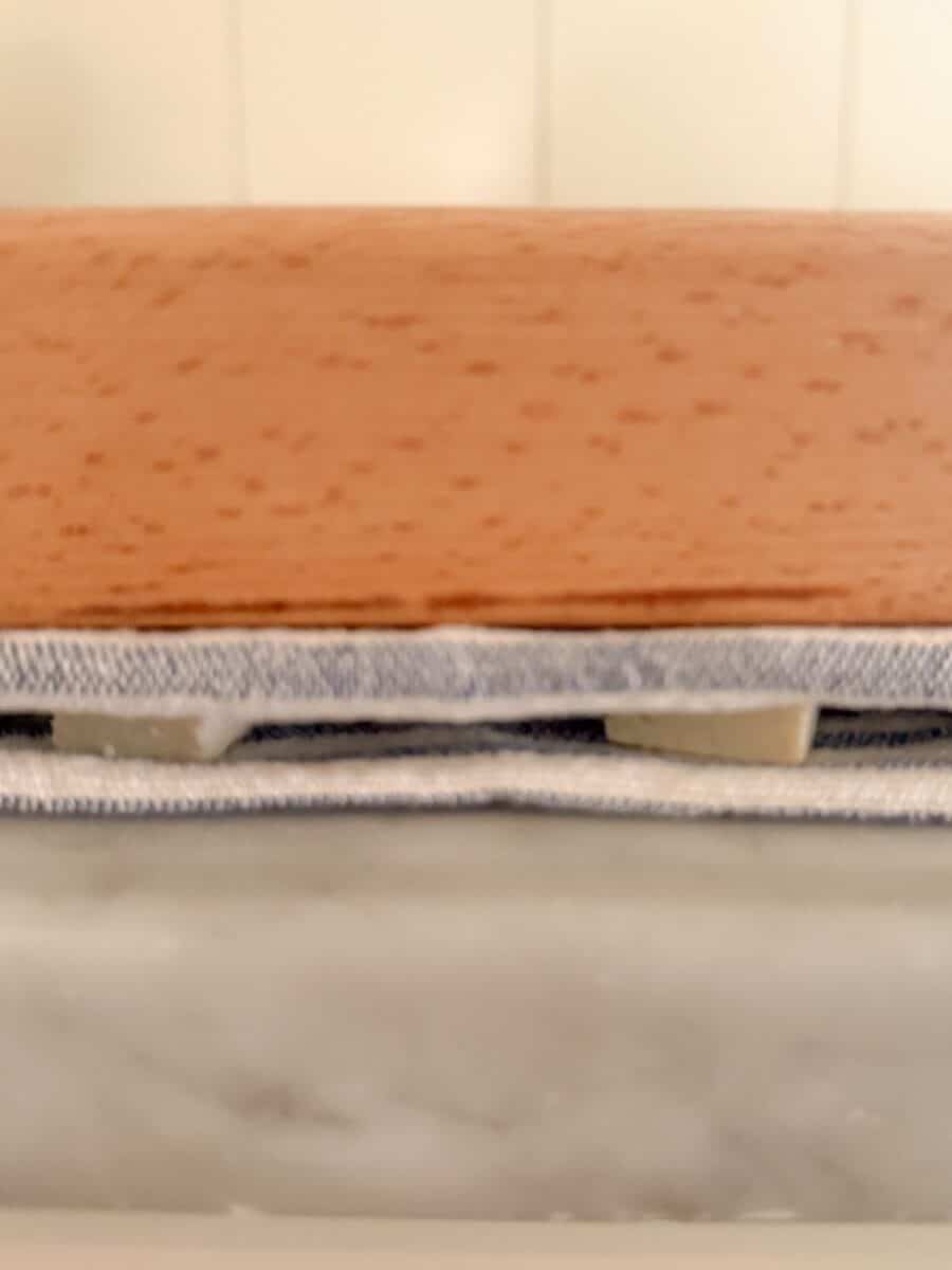 Tofu slices sandwiched between two dishtowels with a cutting board resting on top to press the liquid from the tofu.