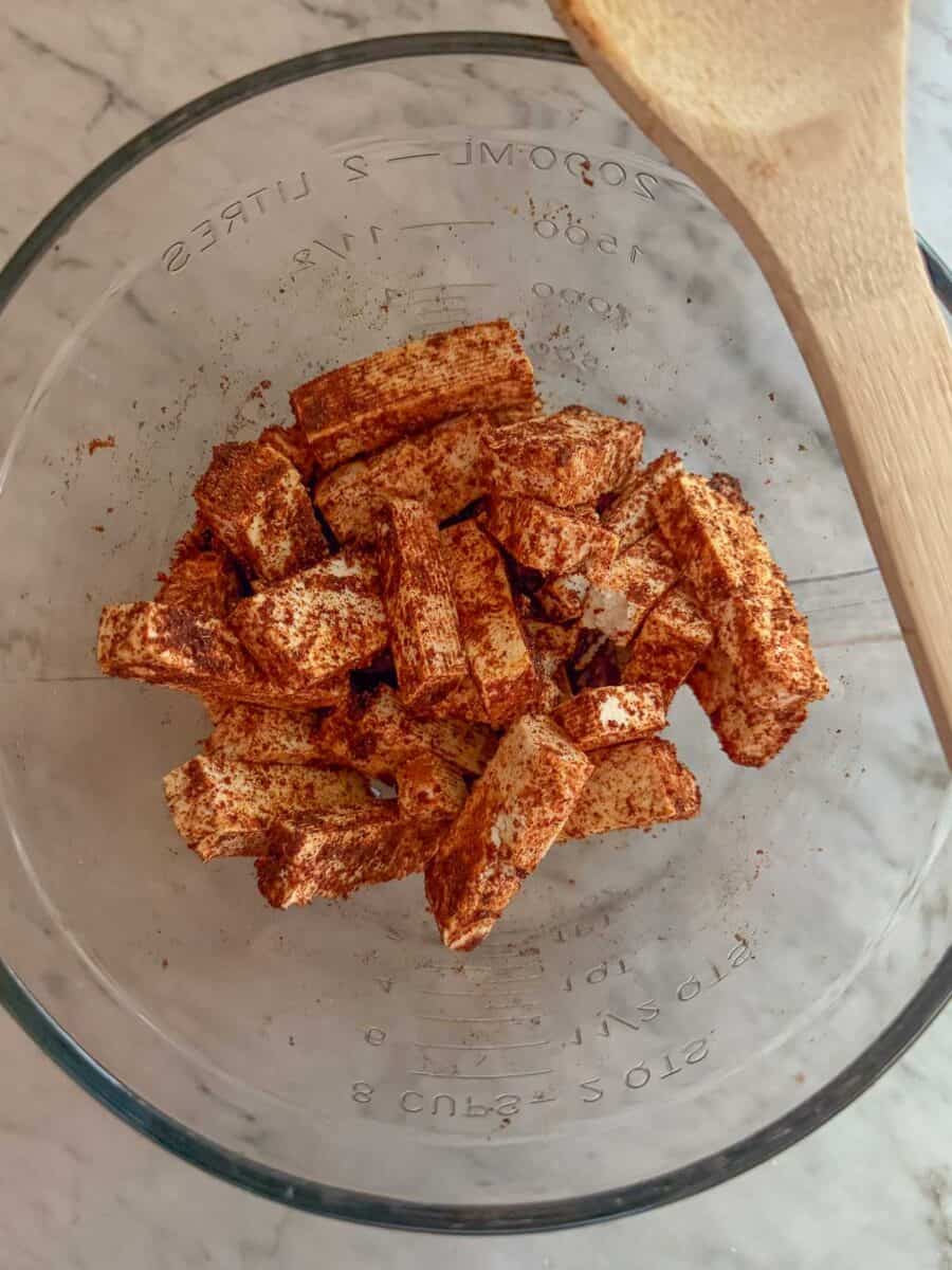 Tofu slices with EVOO and low sodium fajita seasoning on them in a glass mixing bowl with a wooden spoon resting on it.