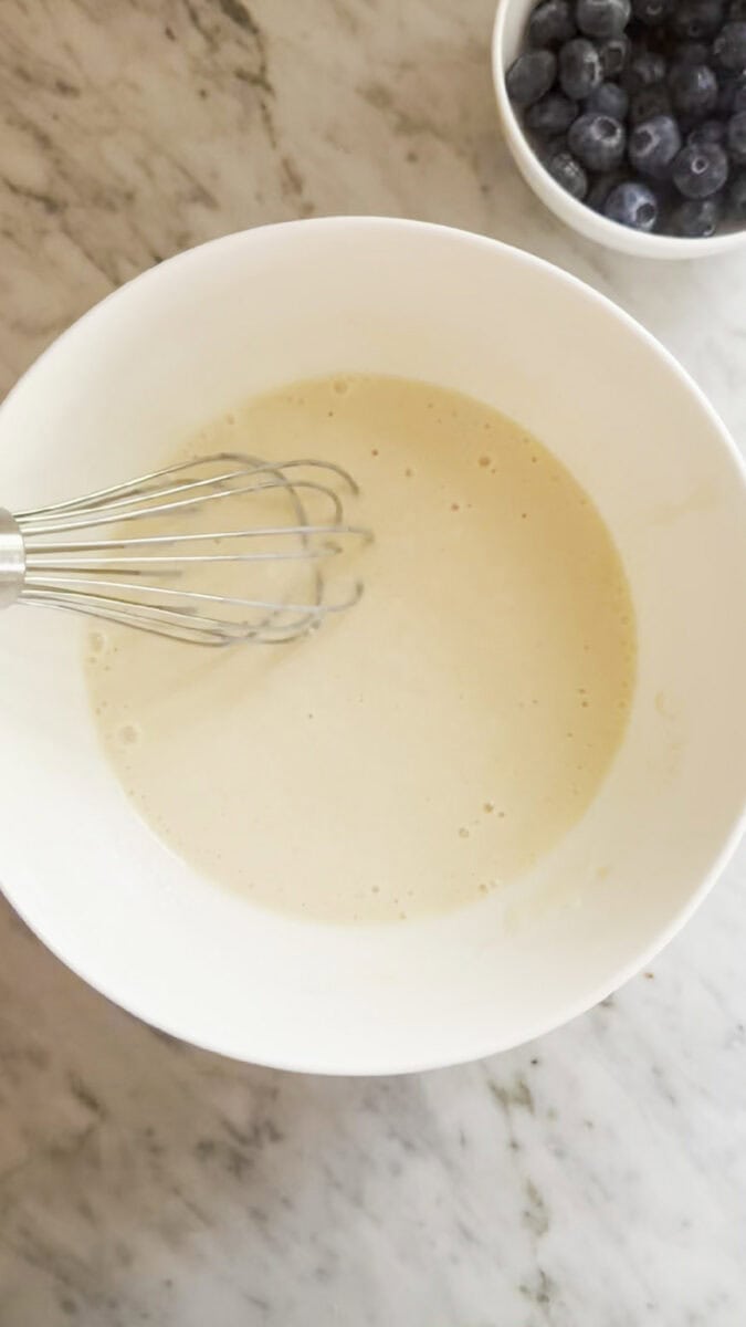 Wet ingredients mixed in a bowl with a whisk. Blueberries are in a small bowl next to it.