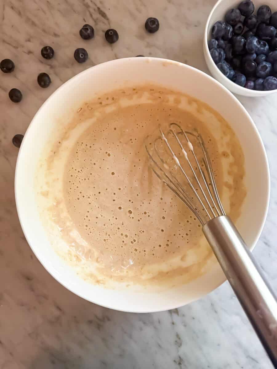 Blueberry protein pancake batter in a white bowl with a whisk on a countertop. Blueberries are in a small bowl next to it.