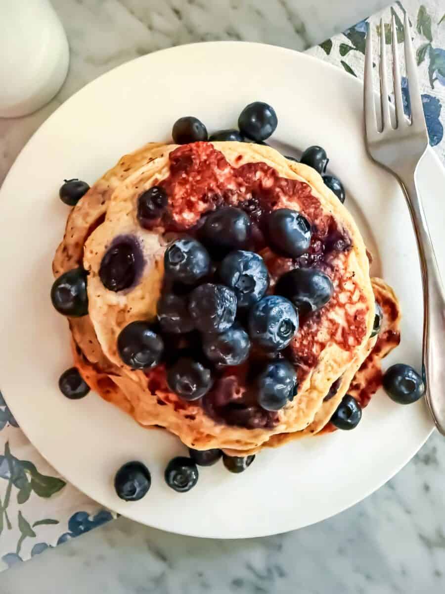 Blueberry protein pancakes topped with fresh blueberries on a white plate with a fork. A cup of coffee is next to it. They are placed on a blueberry printed dish towel on a marble countertop.