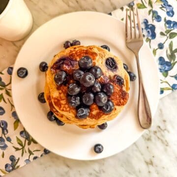 Blueberry protein pancakes topped with fresh blueberries on a white plate with a fork. A cup of coffee is next to it. They are placed on a blueberry printed dish towel on a marble countertop.