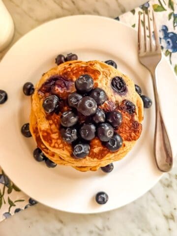 Blueberry protein pancakes topped with fresh blueberries on a white plate with a fork. A cup of coffee is next to it. They are placed on a blueberry printed dish towel on a marble countertop.