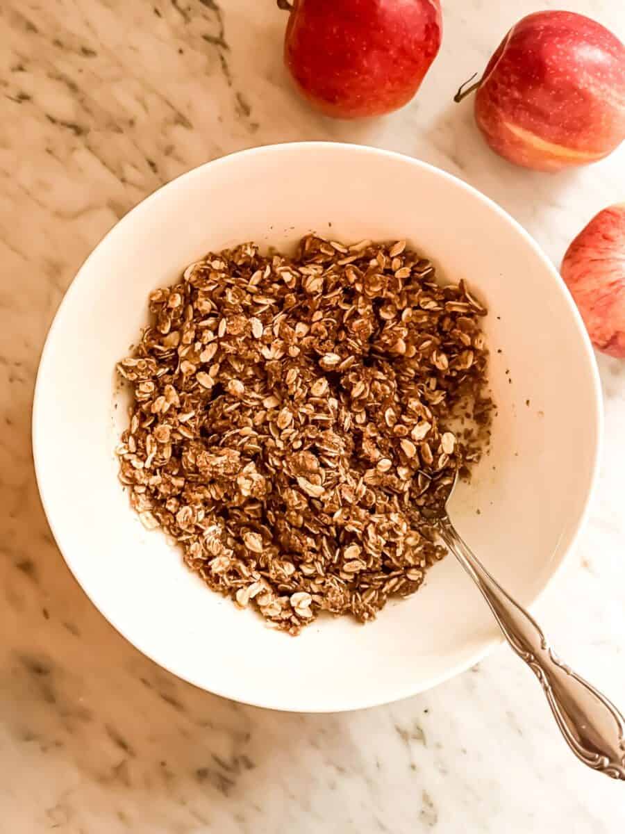 Cherry apple crisp topping in a white bowl surrounded by apples on a marble countertop.