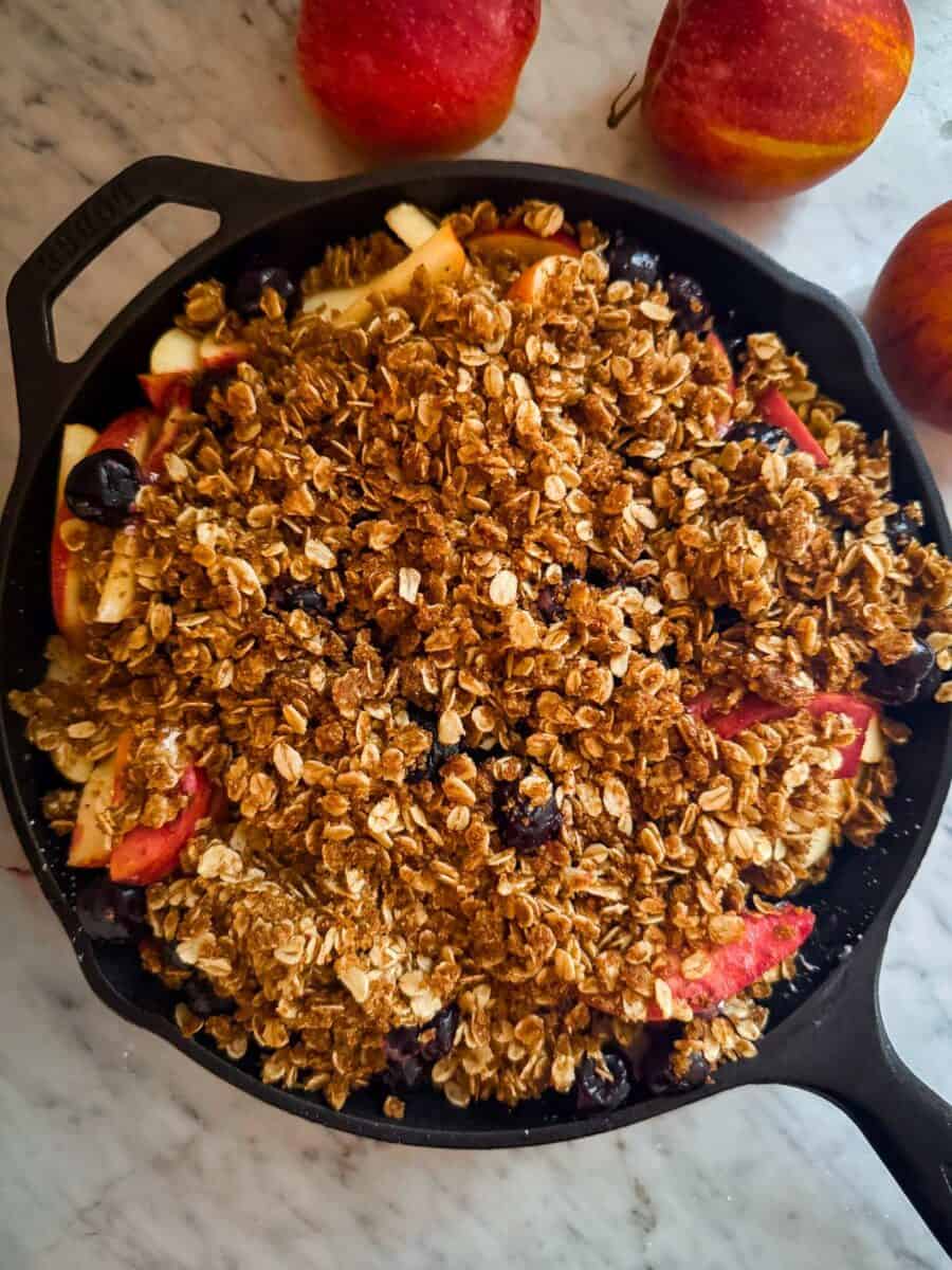 Uncooked cherry apple crisp in a cast iron skillet surrounded by apples on a marble counter.