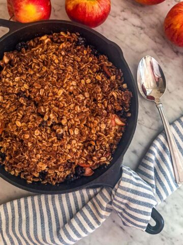 Cherry apple crisp in a cast iron skillet surrounded by apples and a dish towel on a marble counter.