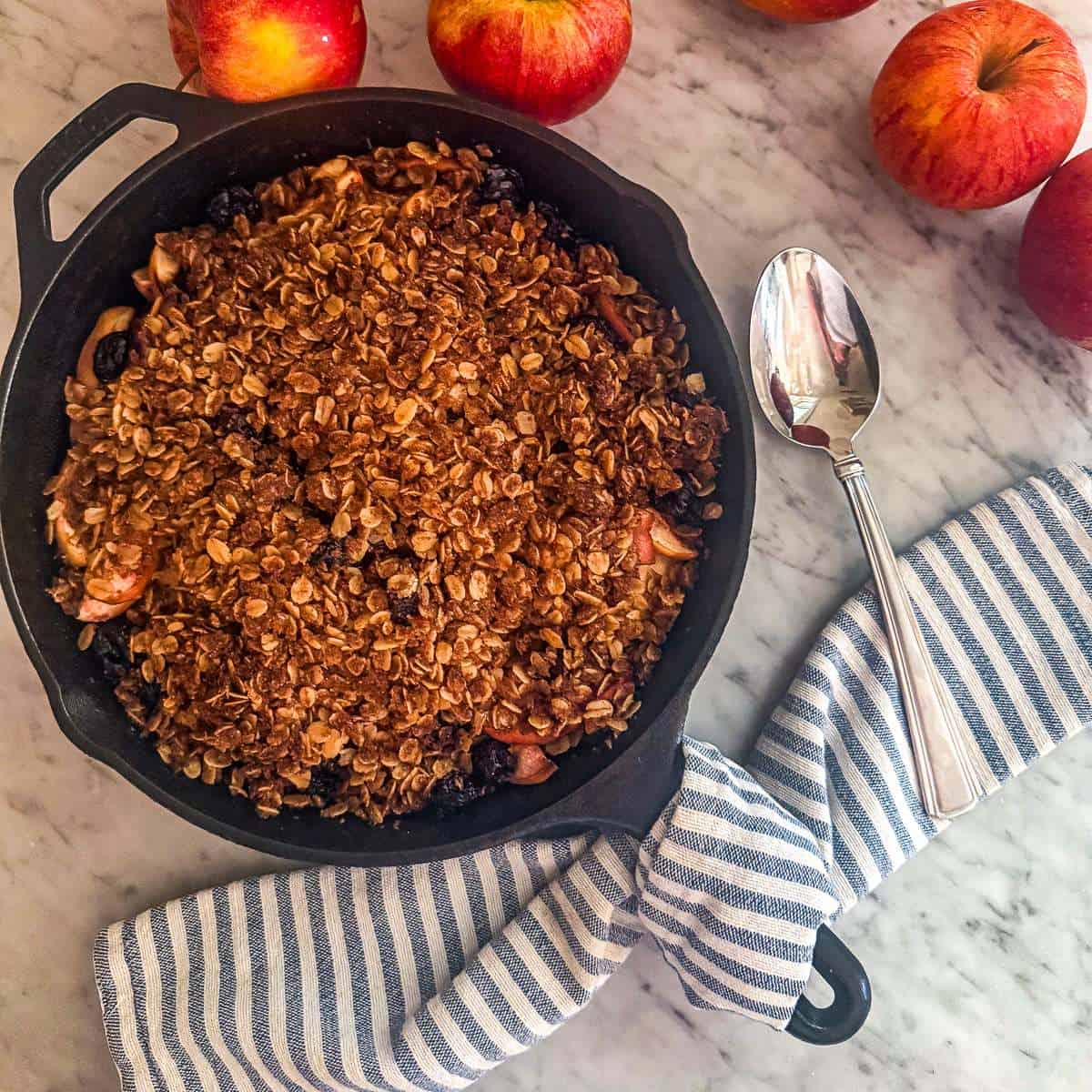 Cherry apple crisp in a cast iron skillet surrounded by apples and a dish towel on a marble counter.