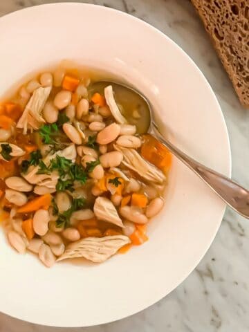 Chicken bean soup prepared in a bowl and garnished with parsley. Whole wheat sourdough bread is to the side.