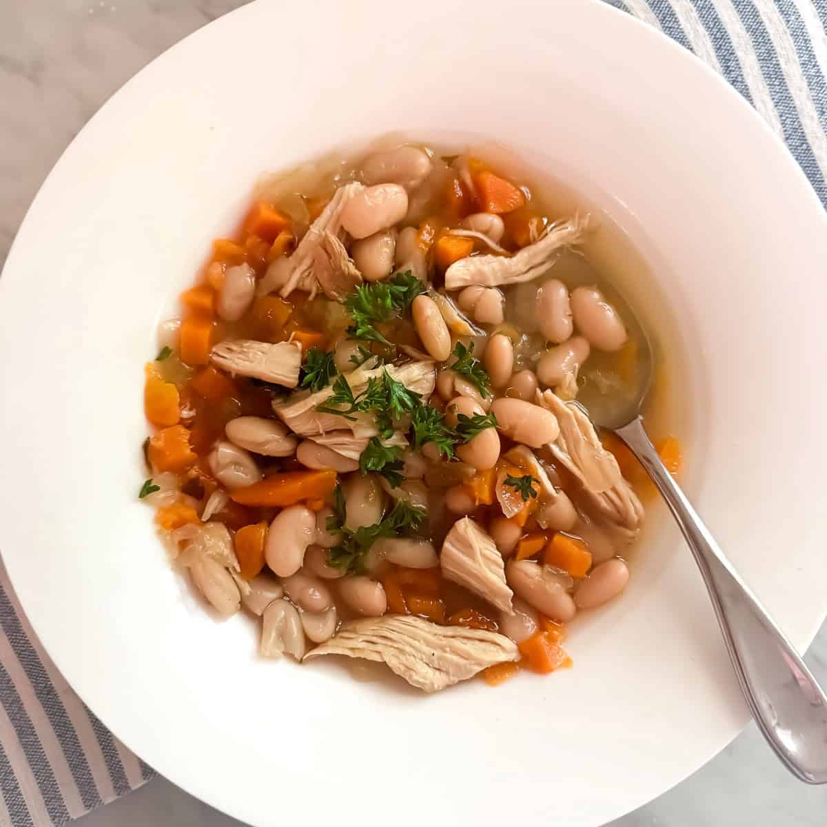 Chicken bean soup prepared in a bowl and garnished with parsley. The bowl is resting on a clue and white dish towel.