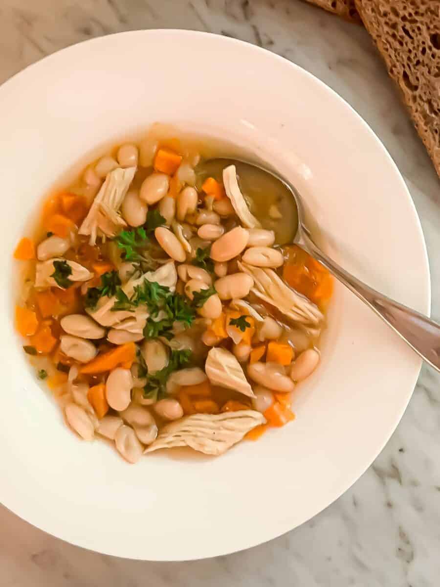 Chicken bean soup prepared in a bowl and garnished with parsley. Whole wheat sourdough bread is to the side.