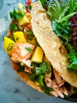 Taco with mango jalapeno salsa, salmon, and black beans with a side salad on a black plate resting on a marble countertop. A glass of water is next to it.