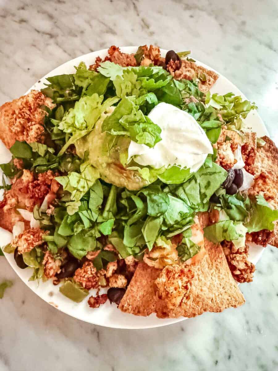 A plate of whole wheat tortilla chips topped with tofu crumbles, Greek yogurt, guacamole, lettuce, cilantro and black beans.