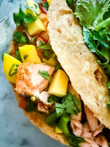 Taco with mango jalapeno salsa, salmon, and black beans with a side salad on a black plate resting on a marble countertop. A glass of water is next to it.