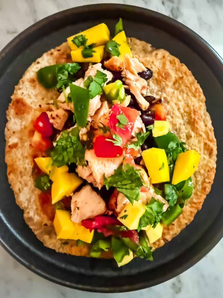 A black plate with a tortilla open flat on it with black beans, mango salsa, and salmon on it. The plate is resting on a marble countertop.