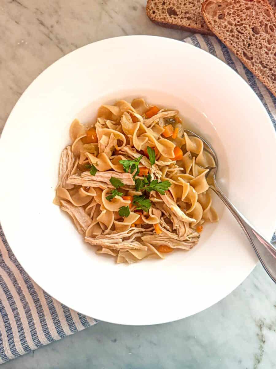 Savory Mediterranean diet chicken noodle soup with vegetables and fresh herbs in a white bowl on a marble surface.