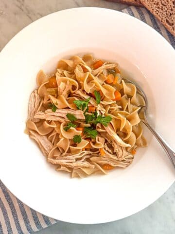 Savory Mediterranean diet chicken noodle soup with vegetables and fresh herbs in a white bowl on a marble surface.
